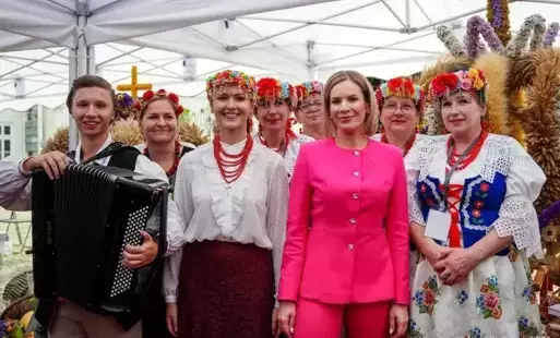 Grupa uczestniczek konkursu wieńcowego w strojach ludowych wraz z akordeonistą i Pierwszą Damą Martą Nawrocką, stojących wspólnie na tle dożynkowych wieńców podczas Dożynek Prezydenckich