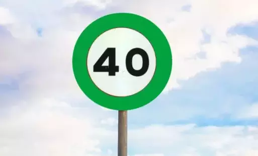 Znak drogowy o okrągłym kształcie z zieloną obwódką i białym tłem, na którym widnieje liczba '40' w czarnym kolorze. Znak umieszczony jest na metalowym słupku, a w tle widać niebo z białymi chmurami