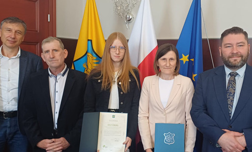 w tle flaga Woźnik, Polski i UE, przed flagami stoją dyrektor LO, Jacek Cogiel, Julia Cogiel, Iwona Cogiel i burmistrz Woźnik Michał Aloszko. 
